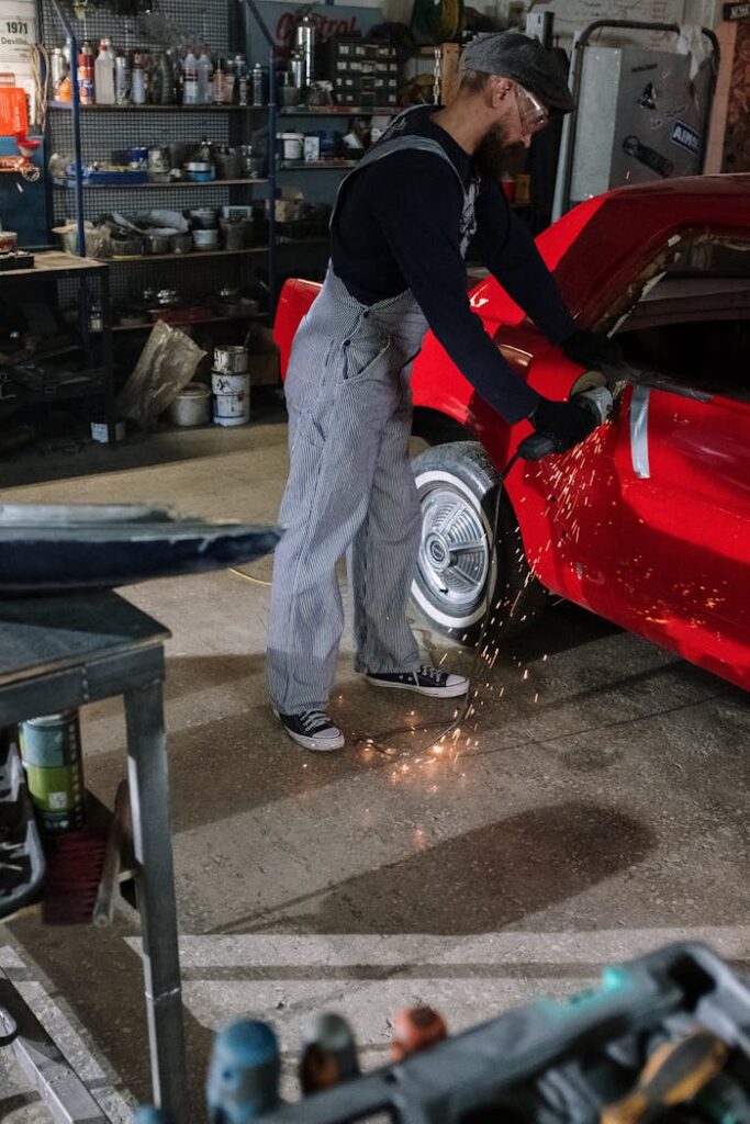 An auto mechanic using a grinder on a red car, creating sparks in a garage setting.